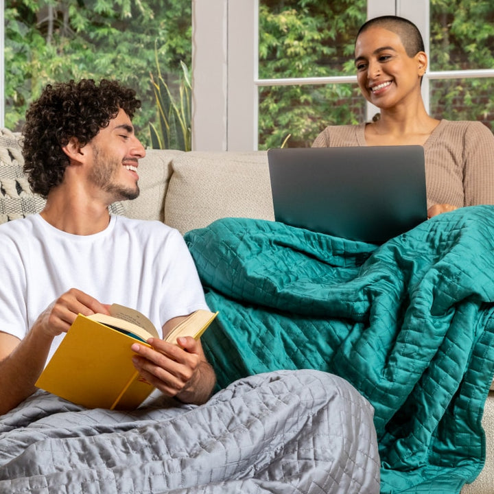 A happy couple sitting on a sofa each with a Gravity Weighted Cooling Blanket - #color_forest-green