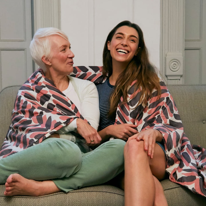 Older woman and younger woman sitting next to each other on a couch covered by a pink and brown blanket