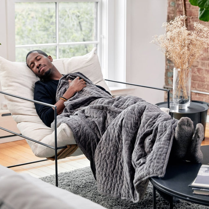 Man covered by a grey blanket lounging on a white chair with his feet up on a coffee table - #color_grey