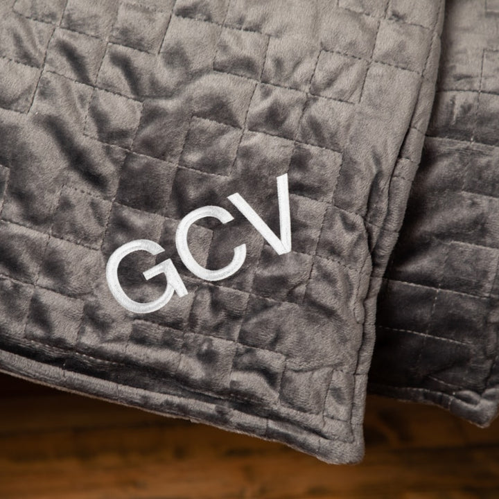 Closeup of the corner of a grey blanket draped over couch embroidered with the initials GCV - #color_grey