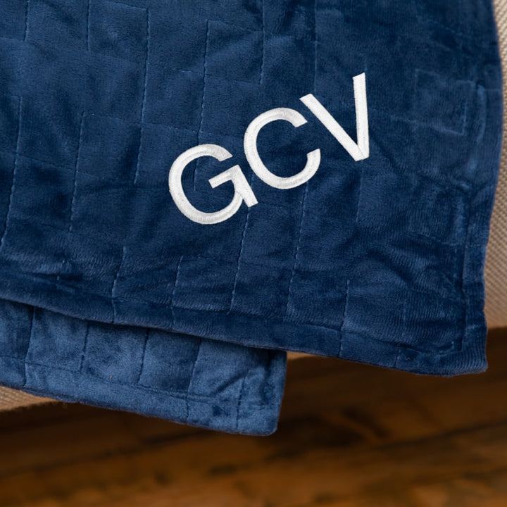 Closeup of the corner of a navy blanket draped over couch embroidered with the initials GCV - #color_navy