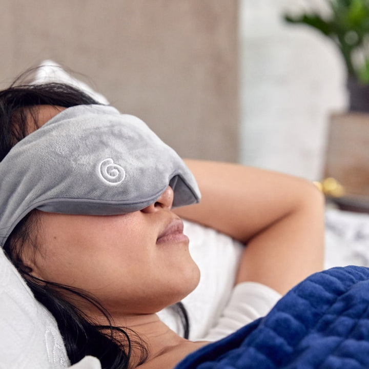 Closeup of woman's face in bed wearing a plush grey weighted eye mask