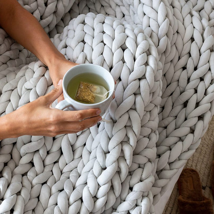 ASoft Grey Gravity Weighted Chunky Knit Blanket on a woman's lap her hands holding a cup of tea over the blanket - #color_soft-grey
