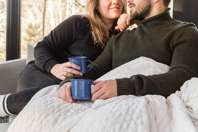 a couple sitting on a couch with mugs and a weighted blanket