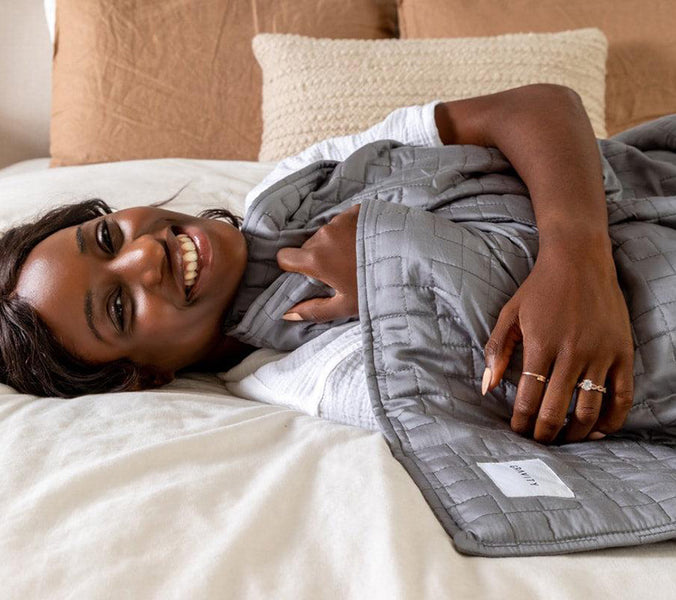 a smiling woman laying under a grey weighted blanket